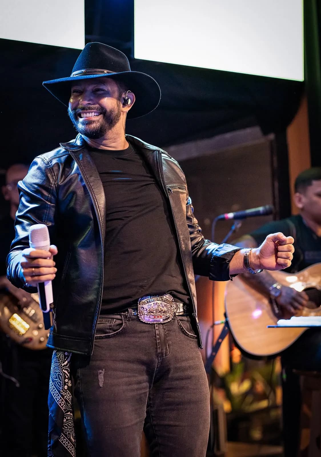 Lukas Santos sorrindo de chapéu, com a banda ao fundo